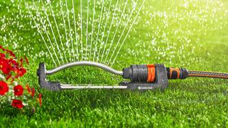 Das Bild zeigt einen dunkelgrau-orangen Rasenspränkler von Garden, welcher Wasser sprüht und rechts der Schlauch angesteckt ist auf einer grünen Wiese.