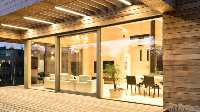 Das Bild zeigt eine moderne Terrasse aus Holz mit überdachtem Bereich und eingebauter Beleuchtung. Große Glasfronten geben den Blick in einen stilvollen, hell beleuchteten Wohn- und Essbereich frei. Im Hintergrund sind weitere Häuser und Bäume zu sehen.