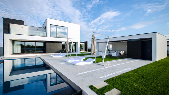Das Bild zeigt eine moderne Luxusvilla mit einem minimalistischen, kubischen Design in Weiß und Schwarz. Die Villa verfügt über große Fensterfronten, eine weitläufige Terrasse mit Sonnenliegen, Sonnenschirmen und einen Pool. Die gepflegte Rasenfläche und der blaue Himmel unterstreichen die luxuriöse und offene Atmosphäre des Anwesens.