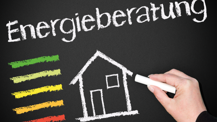 Das Bild zeigt das schwarz einer Kreidetafel, wobei man eine Hand mit Kreide sieht. Auf der Tafel steht bereits "Energieberatung", es wurde ein einfaches Haus gezeichnet und links im Bild sind die Farben für den Energieverbrauch bzw. -stärke angezeichnet. Von grün, über gelb bis rot.