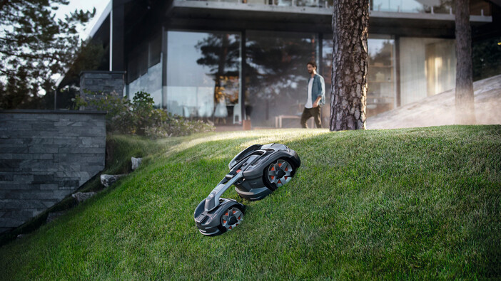 Auf dem Bild ist ein modernes Haus mit einer großen Glasfront zu sehen, das sich in einer bewaldeten Umgebung befindet. Im Vordergrund mäht ein futuristisch aussehender, autonomer Rasenmähroboter eine steile Rasenfläche. Eine Person läuft im Hintergrund nahe dem Haus. Das Bild vermittelt eine High-Tech-Atmosphäre mit Fokus auf smartes Wohnen und automatische Gartenpflege.