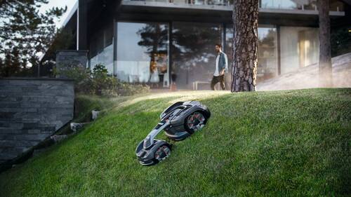 Auf dem Bild ist ein modernes Haus mit einer großen Glasfront zu sehen, das sich in einer bewaldeten Umgebung befindet. Im Vordergrund mäht ein futuristisch aussehender, autonomer Rasenmähroboter eine steile Rasenfläche. Eine Person läuft im Hintergrund nahe dem Haus. Das Bild vermittelt eine High-Tech-Atmosphäre mit Fokus auf smartes Wohnen und automatische Gartenpflege.