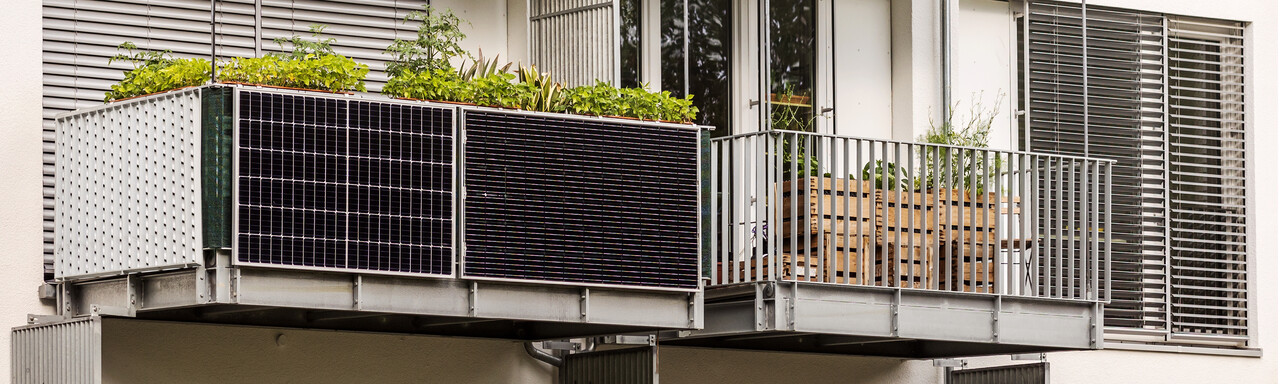 „Ein moderner Balkon mit Solarpanelen und Pflanzenkästen. Der Balkon ist mit grünen Pflanzen und Kräutern geschmückt, während eine Reihe von schwarzen Solarpanelen die Wand entlang verläuft. Der Raum wirkt nachhaltig und umweltbewusst, mit modernen Metallgittern und einer hellen Fassade im Hintergrund.“