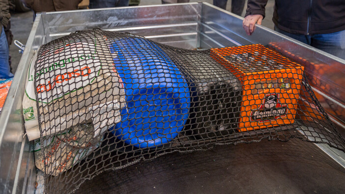 Auf dem Bild ist die Ladefläche eines Anhängers zu sehen, auf der verschiedene Gegenstände mit einem schwarzen Sicherheitsnetz befestigt sind. Darunter befinden sich ein blauer Plastikkanister, eine orange Getränkekiste und ein Sack mit einer Aufschrift, die auf Futtermittel oder Baustoff hindeuten könnte. Um den Anhänger herum stehen Personen, von denen nur teilweise Hände und Kleidung sichtbar sind.