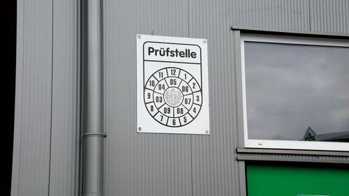 Das Bild zeigt die Blechfassade eines grauen Gebäudes. Links ist eine graue Regenrinne, rechts ein Fenster mit weißen Rahmen und darunter eine grüne Tür mit dem Schriftzug "Notausgang freihalten" zu sehen. In der Mitte befindet sich ein weißes Schild mit der Aufschrift "Prüfstelle" und darunter befinden sich die Zahlen von eins bis zwölf kreisförmig und im Uhrzeigersinn angeordnet. Innerhalb dieses Kreises befindet sich noch ein Kreis mit den Zahlen von Drei bis Neun mit einem Nuller vorgestellt, gegen den Uhrzeigersinn und der Dreier fängt links unten an. Innerhalb dieses Kreises befindet sich noch die Abbildung eines Adlers.