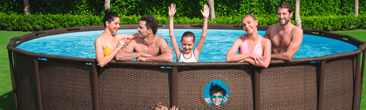 Auf dem Bild ist ein großer runder brauner Pool mit Sichtfenster in einem Garten zu sehen. Sechs Personen befinden sich im Pool, wobei ein Junge durch das Sichtfenster hinaus schaut und ein Mädchen hockelt vor dem Pool. Im Hintergrund sind 5 Bäume, eine große lange Strauchwand und dadurch Hausdächer zu sehen.