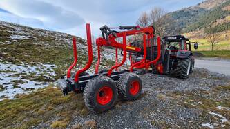 Das Bild zeigt einen schwarzen Holztransporter mit roten Felgen und Holzhalterungen und großen schwarzen Reifen auf einem Hügel mit grüner Wiese und einem Steinweg mit schwarzen Steinen. Im Hintergrund sieht man einen schwarz-roten Traktor von hinten mit roten Palfingerkran, einen einzelnen Baum direkt vor dem Traktor und weiter im Hintergrund noch einen Hügel mit vielen Bäumen. Auf der Wiese liegt noch ein wenig Schnee und der Himmel ist etwas dunkel bewölkt.
