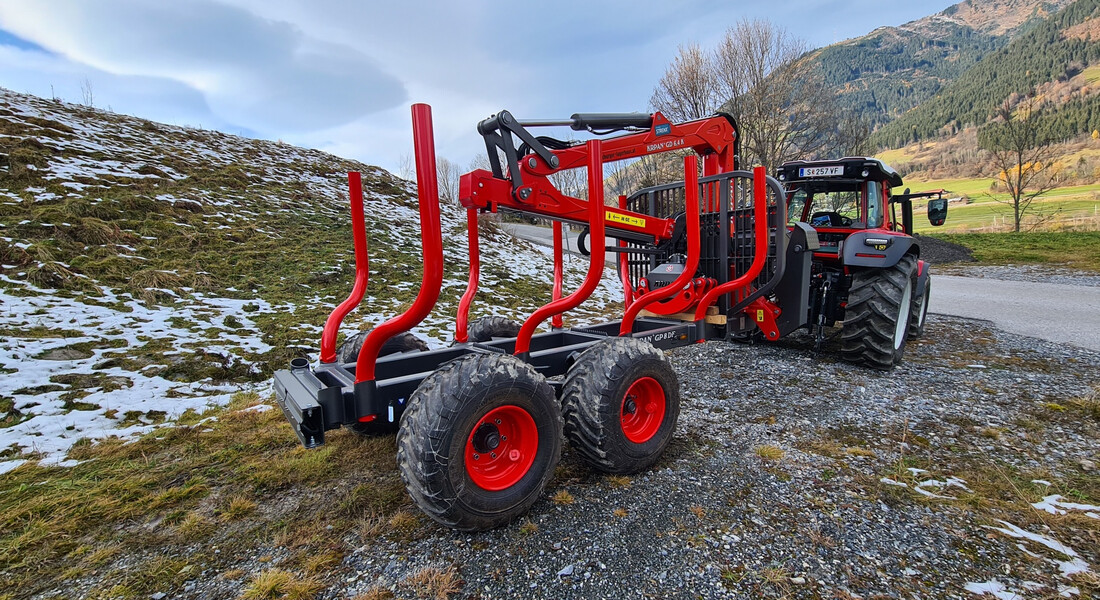 Das Bild zeigt einen schwarzen Holztransporter mit roten Felgen und Holzhalterungen und großen schwarzen Reifen auf einem Hügel mit grüner Wiese und einem Steinweg mit schwarzen Steinen. Im Hintergrund sieht man einen schwarz-roten Traktor von hinten mit roten Palfingerkran, einen einzelnen Baum direkt vor dem Traktor und weiter im Hintergrund noch einen Hügel mit vielen Bäumen. Auf der Wiese liegt noch ein wenig Schnee und der Himmel ist etwas dunkel bewölkt.